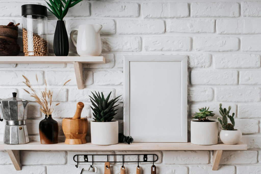 A minimalist kitchen wall with white brick tiles features two wooden shelves. The shelves hold plants in white pots, a mortar and pestle, a coffee maker, a pitcher, and a picture frame. Below are hanging kitchen utensils.