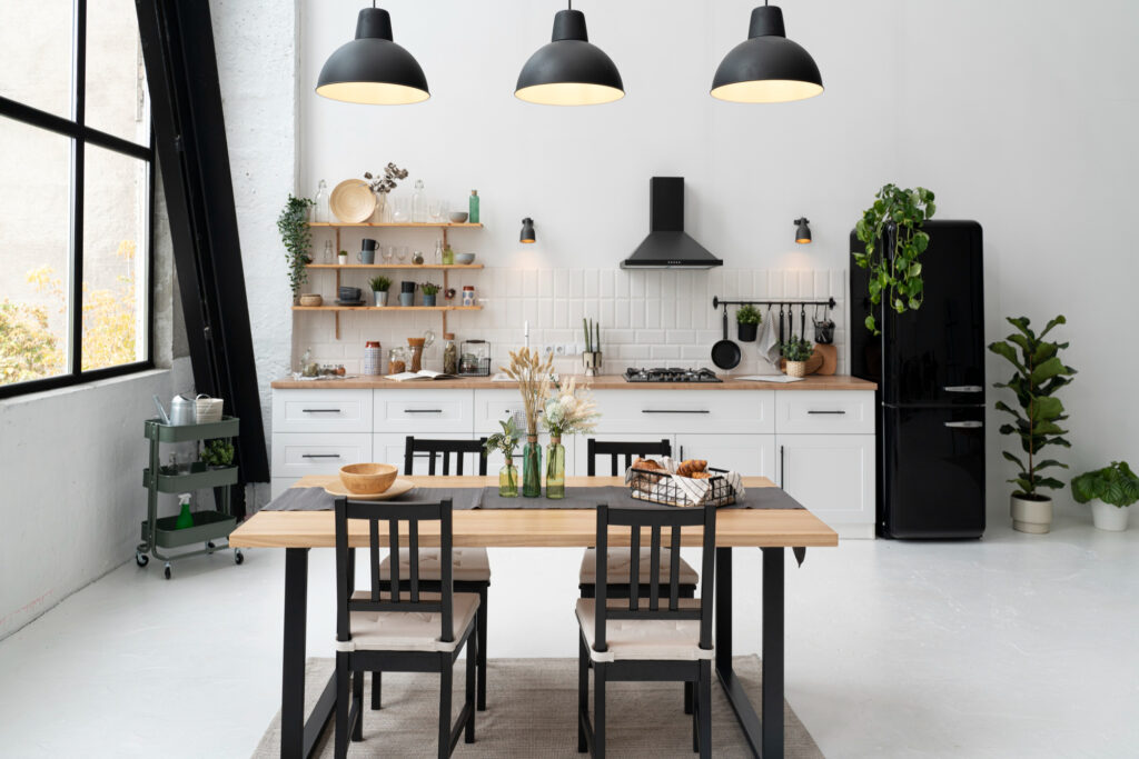 Modern kitchen with a wooden dining table set for four. White cabinetry, open shelves with plants, a black fridge, and three pendant lights overhead. Large window on the left and a potted plant in the corner enhance the airy space.