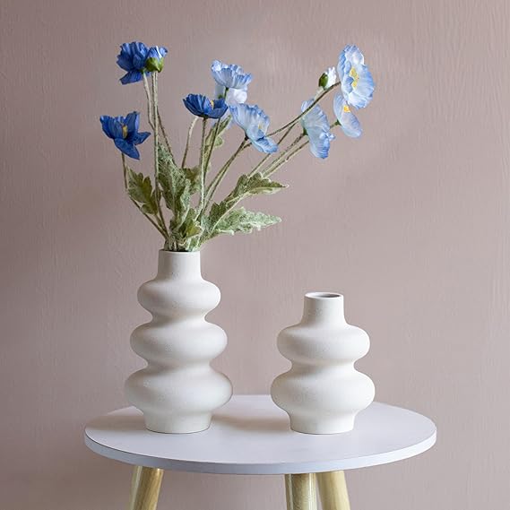 A white, round table holds two cream-colored vases. The taller vase contains blue flowers with green stems, while the shorter vase is empty. The background is a plain, light beige wall.