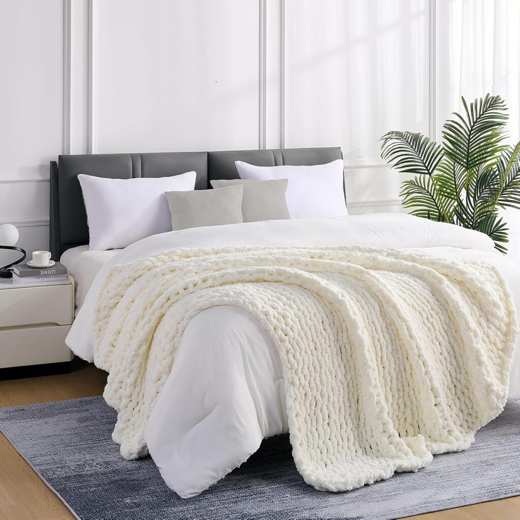 A cozy bedroom featuring a neatly made bed with white linens and a textured cream knit blanket. Two white pillows and a gray cushion rest against a dark headboard. A potted plant stands beside a light-colored nightstand with a cup and book.