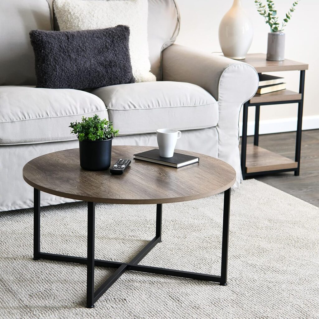 A cozy living room with a light gray sofa, dark cushions, and a round wooden coffee table. The table holds a small potted plant, a cup, a book, and a remote. A side table with a lamp and books is in the background, and a textured rug covers the floor.