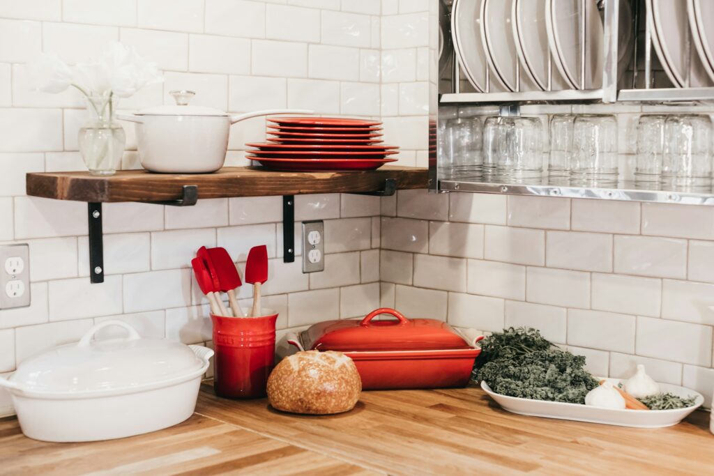 A kitchen corner with white tile walls features a wooden countertop. On it are a white casserole dish, red utensils in a holder, a red dish, a loaf of bread, kale, and onions. A shelf holds stacked red plates and white bowls, with glasses below.