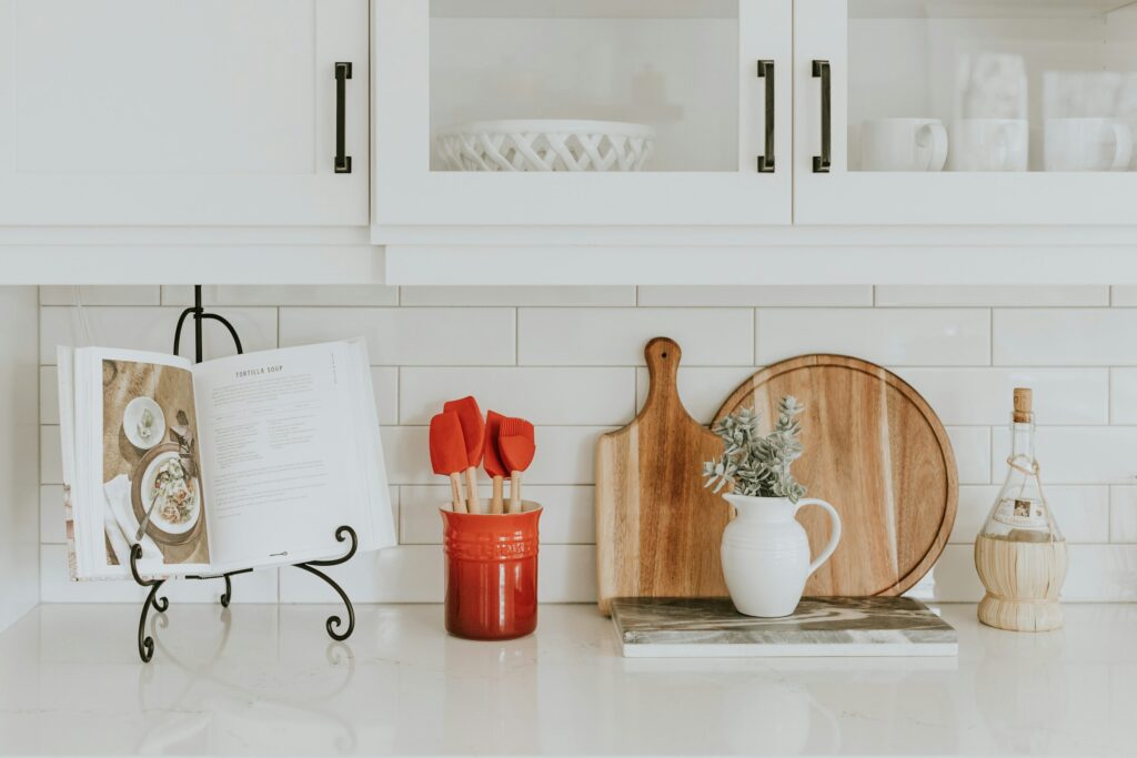 A kitchen countertop with a cookbook on a stand, a red container holding red spatulas, a wooden cutting board, a round wooden tray, a white pitcher with green foliage, and a decorative bottle, against a white tile backsplash and cabinets.