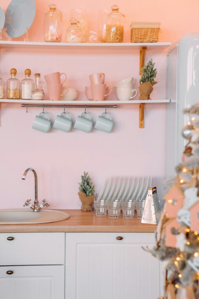 A pastel-themed kitchen with pink and blue mugs hanging. Shelves display jars and dishes. A tree with lights is partially visible on the right. A white sink, faucet, and wooden countertop complete the cozy scene.