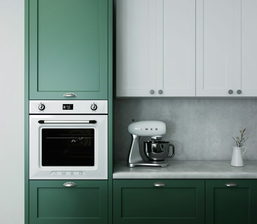 A modern kitchen with green and white cabinetry. Theres a built-in oven and a white stand mixer on the countertop. A small white jug with a twig is also on the countertop, set against a gray backsplash.