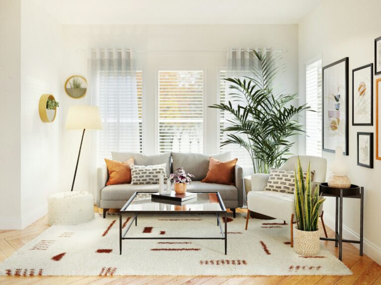 A cozy living room with a gray sofa, white armchair, and a patterned rug. A floor lamp stands beside the sofa. Various plants decorate the space, with wall art and a large window allowing natural light to brighten the room.